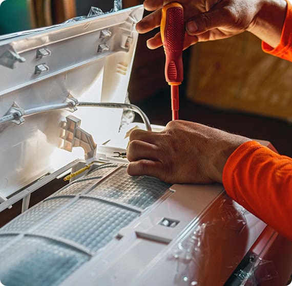 Osoba w pomarańczowej koszuli z długim rękawem używa śrubokręta do naprawy lub instalacji jednostki klimatyzacyjnej. Pokrywa jednostki jest otwarta, odsłaniając wewnętrzne komponenty i okablowanie.